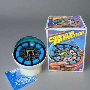 May include: A vintage Computer Perfection electronic game featuring a white base, a circular black control panel with blue buttons, and a translucent teal dome cover. The set includes the original cardboard box and a printed instruction manual.