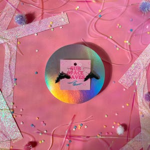 May include: A pair of black bat-shaped earrings on a pink card with the text "SUB SPACE AVES". The earrings are displayed on a holographic circle, surrounded by pink confetti and glittery decorations.