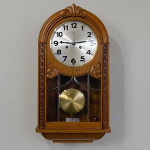 May include: A wall clock with a brown wooden frame featuring carved details, an arched top, and a glass-fronted lower section. The clock face is silver with black Arabic numerals from 1 to 12 and smaller numbers 13 to 24. A gold-coloured pendulum is visible.
