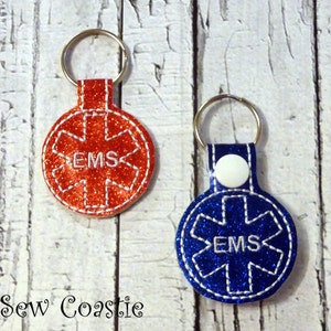 Puede incluir: Dos llaveros con un diseño de estrella de la vida roja y azul. Los llaveros están hechos de tela de brillantina y tienen las letras "EMS" bordadas en blanco.