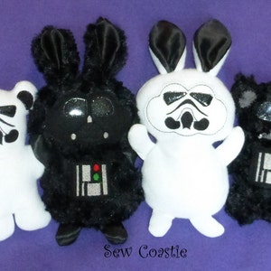 May include: Four plush toys, each with a different design inspired by Star Wars characters. The toys are a white bear with a Stormtrooper helmet, a black bunny with a Darth Vader helmet, a white bunny with a Stormtrooper helmet, and a black bear with a Darth Vader helmet. The toys are all standing on a purple surface.