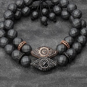 May include: A black beaded bracelet with two evil eye charms. The charms are made of rose gold and black stones.