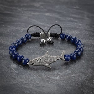 Puede incluir: Un brazalete de cuentas de lapislázuli azul con un dije de tiburón de plata. El brazalete tiene un cordón negro con un cierre de palanca de plata.