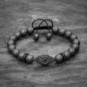 May include: Black beaded bracelet with a black evil eye charm. The bracelet is made of faceted beads and has a black cord closure. Juniper & Eloise Jewelry.