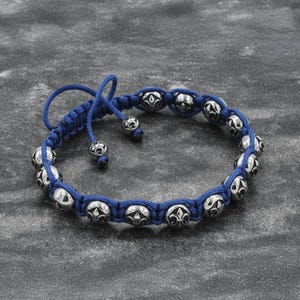 May include: A blue macrame bracelet with silver-colored beads. The beads have a fleur-de-lis design. The bracelet is adjustable with a sliding knot closure. The beads are approximately 0.5 inches in diameter.