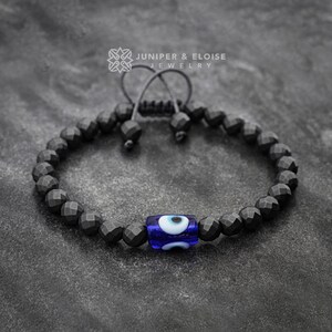 May include: Black beaded bracelet with a blue evil eye charm. The bracelet is made of faceted black beads and has a simple adjustable knot closure. Juniper & Eloise Jewelry.