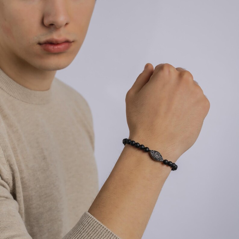 May include: A black beaded bracelet with a black and silver eye charm. The bracelet is on a person's wrist.