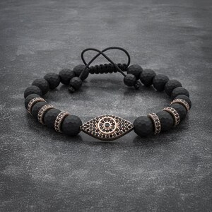 May include: Black beaded bracelet with a gold evil eye charm. The charm is encrusted with black stones.