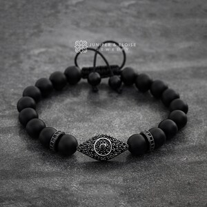 May include: Black beaded bracelet with a black evil eye charm featuring a black crystal center. The bracelet has a black cord closure with a silver toggle.