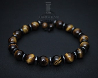 May include: A bracelet made of tiger's eye and dark grey hematite beads. The tiger's eye beads are brown and gold, and the hematite beads are metallic grey. The bracelet is strung on a black cord. The brand name is Juniper & Eloise.