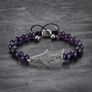 Puede incluir: Una pulsera de cuentas con cuentas de amatista y un dije de tiburón plateado. La pulsera tiene un cierre de cordón ajustable negro y dos cuentas plateadas. El dije de tiburón tiene una textura detallada.