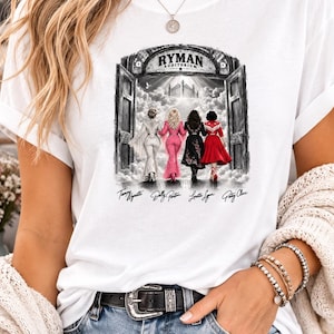 Puede incluir: Una camiseta blanca con un estampado gráfico de cuatro figuras caminando hacia la entrada del Ryman Auditorium. El texto dice Ryman Auditorium e incluye los nombres Tammy Wynette, Dolly Parton, Loretta Lynn y Patsy Cline bajo las figuras.