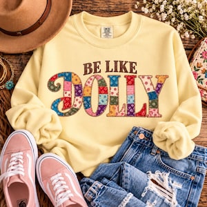 May include: A pale yellow crewneck sweatshirt featuring the brown text BE LIKE and the word DOLLY in large, multicolored patchwork-style letters with various floral and geometric patterns. The garment has a Comfort Colors brand label.