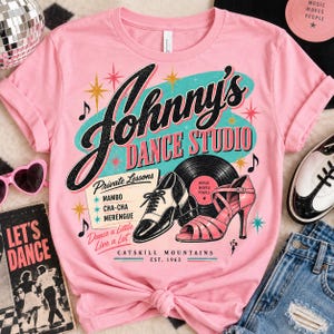 May include: A light pink t-shirt featuring a graphic print that reads Johnny's Dance Studio. The design includes a black and white oxford shoe, a pink high-heeled sandal, a vinyl record, and text listing Private Lessons for Mambo, Cha-Cha, and Merengue.