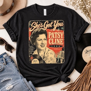 May include: A black t-shirt featuring a vintage-style graphic of Patsy Cline. The print includes the text She's Got You, Patsy Cline, 1962, Nashville Tennessee, and a list of song titles. The design also displays a microphone and a vinyl record.