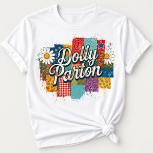 May include: A white short-sleeved t-shirt featuring a central graphic of the text Dolly Parton in a cursive font. The design includes a patchwork-style background with various colorful patterns, including red gingham, floral prints, and a blue star.