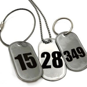 May include: Three silver dog tags with black numbers: 15, 28, and 349. One tag is on a metal loop, another on a ball chain, and the third on a metal ring. The tags are rectangular with rounded edges.