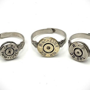 May include: Three silver rings with bullet casings as the centerpieces. The rings are made of metal and have a silver finish. The bullet casings are engraved with the caliber and manufacturer. The first ring has a 9mm Luger casing, the second ring has a 308 Winchester casing, and the third ring has a 38 Special casing.
