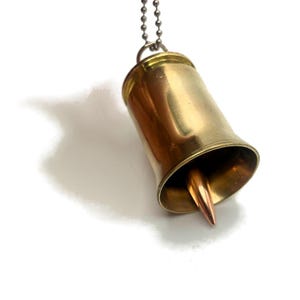 May include: A brass bullet casing pendant with a copper bullet tip hanging from a silver chain. The pendant is shaped like a bell.