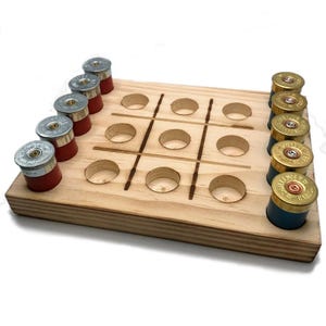 May include: A wooden tic-tac-toe game board with nine holes for playing. The board is made of light wood and has a natural finish. The game is played with shotgun shells, with red and blue shells representing the two players.