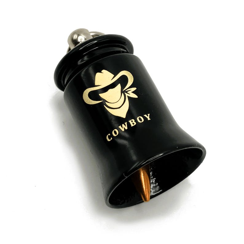 May include: Black bell-shaped pendant with a gold-colored cowboy hat logo and the word "COWBOY" in gold lettering. The pendant has a silver-colored loop at the top and a copper-colored tip. The pendant is likely a keychain or charm.