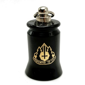 May include: Black bottle stopper with a silver top and ring. The bottle stopper has a gold emblem with a cross, sword, and mountain design, and the words "DEDICATED TO LIFE".