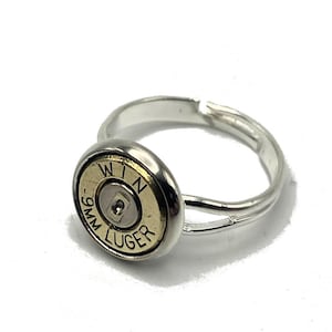 Peut inclure: Bague argentée ornée d'un étui de balle doré en son centre. L'étui porte l'inscription "WIN" au-dessus de "9MM LUGER". La bague est dotée d'un anneau réglable.