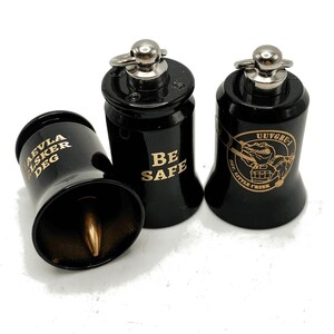 May include: Three black metal bells with silver accents. One bell has the text "AEVLA LSKER DEG" in gold, another reads "BE SAFE", and the third features a cartoon alligator and the text "UUVGRU - MT. LITTLE CREEK" in gold.