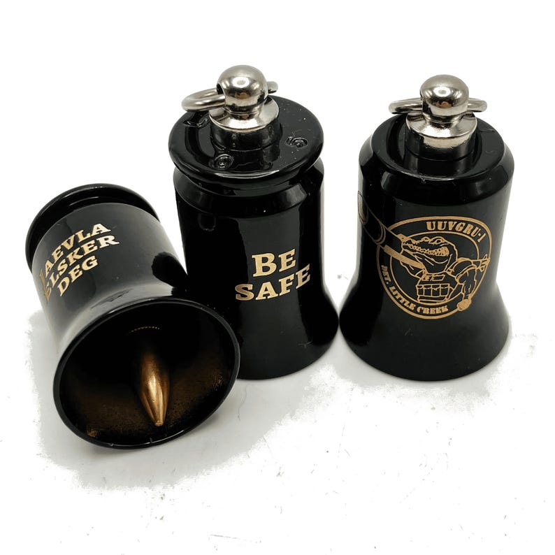 May include: Three black metal bells with silver accents. One bell has the text "AEVLA LSKER DEG" in gold, another reads "BE SAFE", and the third features a cartoon alligator and the text "UUVGRU - MT. LITTLE CREEK" in gold.