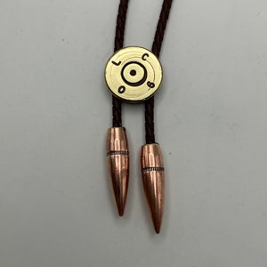 May include: A brown leather bolo tie with a brass bullet casing as the centerpiece. The bullet casing has the letters "LC" and "06" engraved on it. The ends of the tie are made of copper-colored bullet tips.