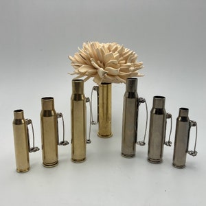 May include: A set of seven bullet casings with pins attached. The casings are made of brass and silver metal. The casings are arranged in a row from largest to smallest.