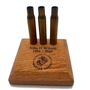 May include: A wooden display stand with three empty bullet casings. The stand is engraved with the text "John H Wilson 1953 - 2024" and the United States Marine Corps logo.