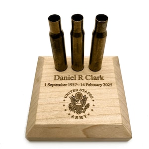 May include: A square light-colored wooden base featuring three upright brass-colored metal casings. The wood is engraved with the name Daniel R Clark, the dates 1 September 1937 to 14 February 2025, and the United States Army emblem.