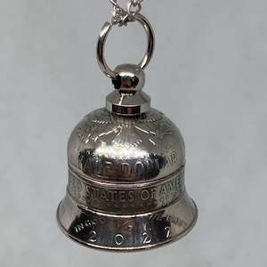 May include: A silver bell pendant with a United States Half Dollar coin design. The coin features an eagle with outstretched wings and the inscription "United States of America" and "Half Dollar". The bell is attached to a silver chain.