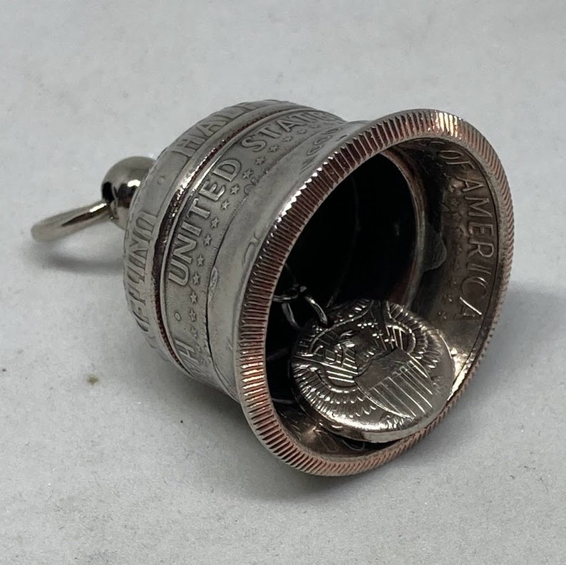 May include: A silver bell made from a United States half dollar coin with a copper rim. The bell has a small silver chain with a silver charm inside. The charm is a United States half dollar coin with the eagle and shield design.
