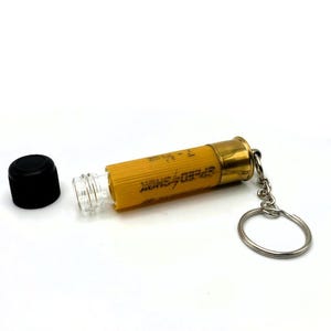 May include: A yellow and brass shotgun shell keychain with the text "SPEED SHOK" printed on the side. The keychain is open, revealing a clear glass interior. A black cap sits next to the shell.