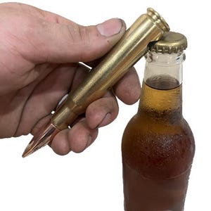 May include: A brass bullet-shaped bottle opener is used to open a brown glass bottle. The bottle opener is held in a hand and is gold in color. The bottle contains a light brown liquid.