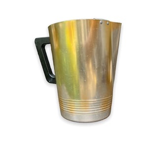May include: A gold metal pitcher with a black plastic handle. The pitcher has a textured surface and a ribbed design around the bottom.