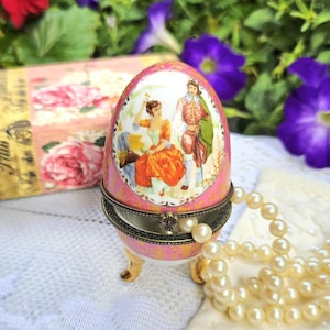 May include: A pink porcelain egg-shaped trinket box with a hinged lid and gold-tone metal feet. The exterior features a central oval illustration of a couple in period clothing, surrounded by gold filigree patterns on a vibrant pink background.