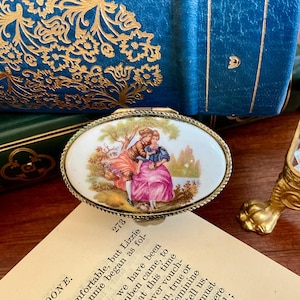 May include: An oval, antique trinket box with a hinged lid. The lid features a painted scene of a couple in a garden. The box has a gold-colored metal frame and is set on a wooden surface. The text "273" is visible.