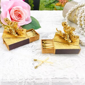 May include: Two rectangular matchboxes with dark brown sides and gold-toned tops, each featuring a small gold-colored cherub figurine. The boxes are partially open, revealing wooden matches inside, with two additional matches lying on the surface.