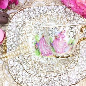 May include: A vintage teacup and saucer set with a gold filigree pattern and a scene of women in pink dresses. The set includes a teacup, a saucer, and a dessert plate. The set is decorated with gold trim and is placed on a lace doily.
