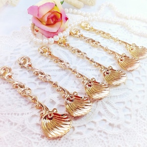 May include: Six ornate gold-colored spoons with seashell-shaped bowls and decorative handles. The spoons are arranged on a white lace surface, with a pink and yellow rose and a pearl necklace in the background. The spoons have a vintage aesthetic.