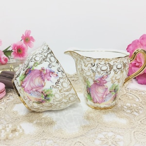 May include: A vintage teacup and creamer set. The white porcelain pieces feature a gold filigree design and a painted scene of a woman in a pink dress. The creamer has a gold handle. Pink flowers and a lace doily are in the background.