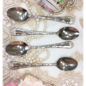 May include: Four silver spoons with ornate floral detailing are arranged on a lace tablecloth, alongside a vintage teacup and a string of pearls. The spoons have a reflective surface, and the scene evokes a sense of vintage elegance.