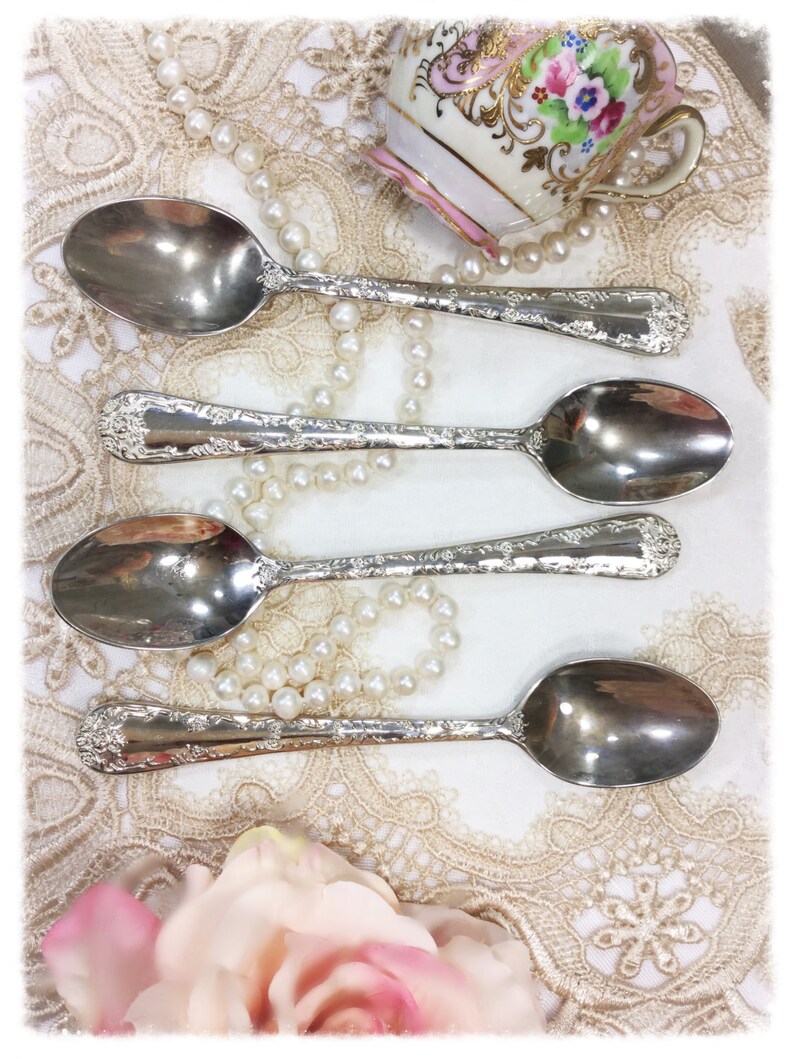 May include: Four silver spoons with ornate floral detailing are arranged on a lace tablecloth, alongside a vintage teacup and a string of pearls. The spoons have a reflective surface, and the scene evokes a sense of vintage elegance.
