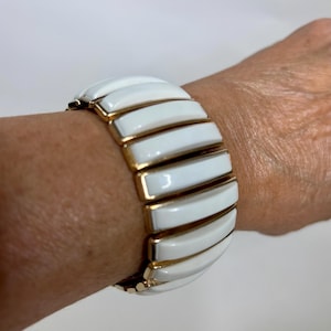 May include: White and gold-tone bracelet. The bracelet features rectangular white enamel segments with gold-tone metal borders. The bracelet is on a wrist. The bracelet is a stretch style.