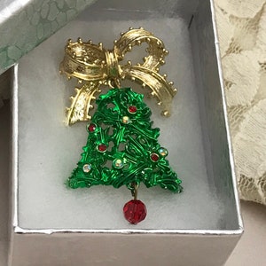 May include: A green Christmas tree-shaped brooch with a gold bow and a red jewel ornament. The brooch is in a white box with a silver lid.