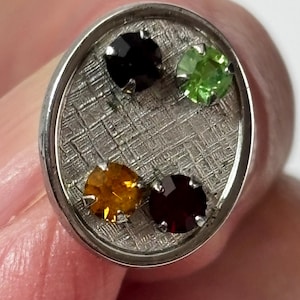May include: Oval silver cufflink with a textured centre, set with four gemstones in black, green, orange, and burgundy. The gemstones are arranged in a circular pattern within the cufflink.