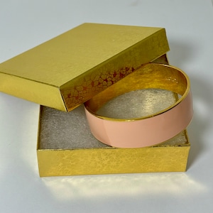 May include: A pink and gold bangle bracelet in a gold gift box. The bangle is wide and has a smooth, glossy finish.